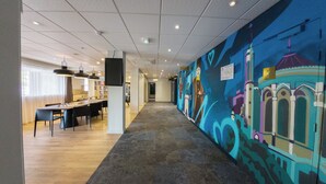 Un couloir orné d’une fresque murale, un espace repas avec des tables et des chaises, et un sol recouvert de moquette.