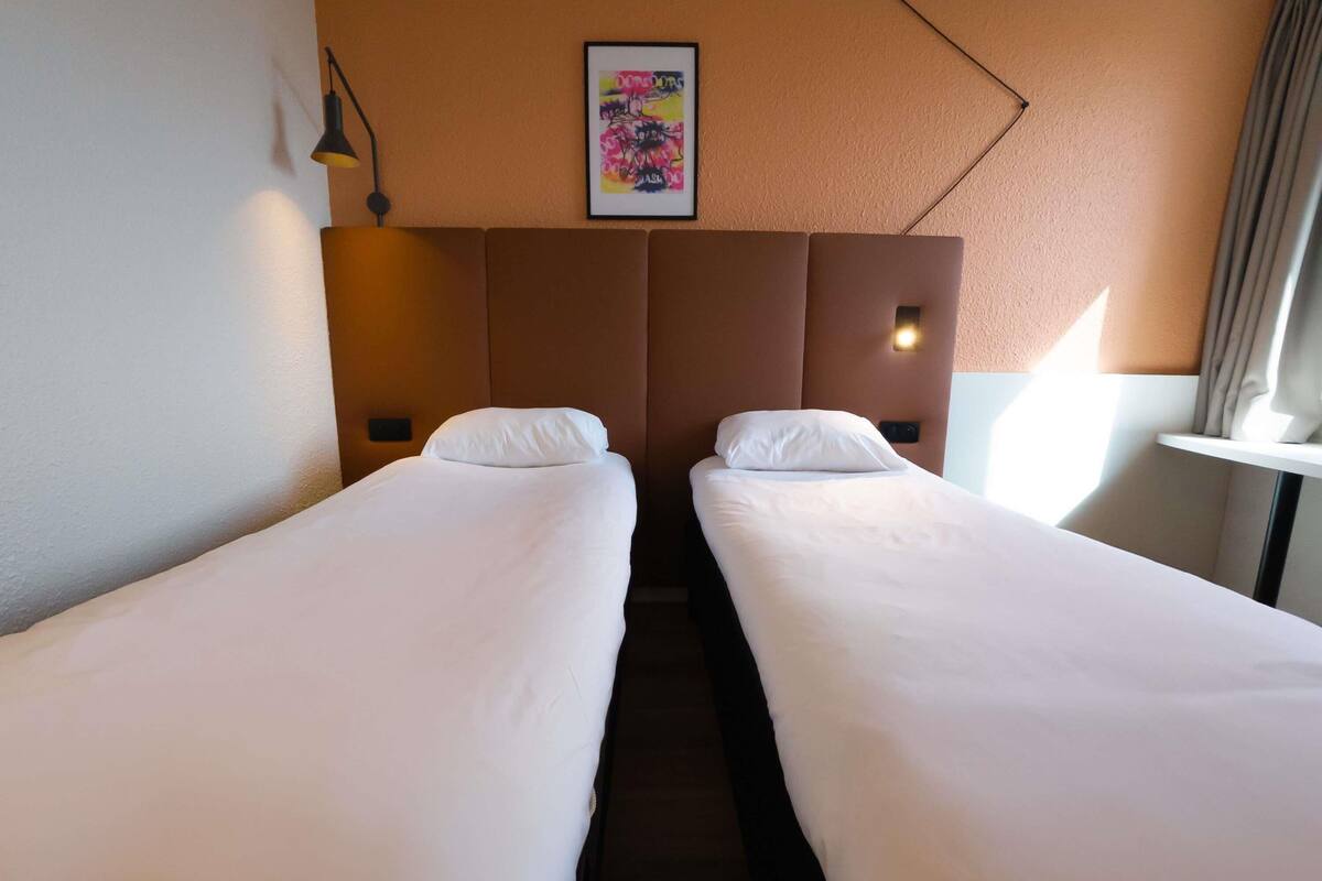 Une chambre d’hôtel avec deux lits simples, une tête de lit dotée d’un luminaire, une œuvre d’art encadrée au mur et une fenêtre avec des rideaux.