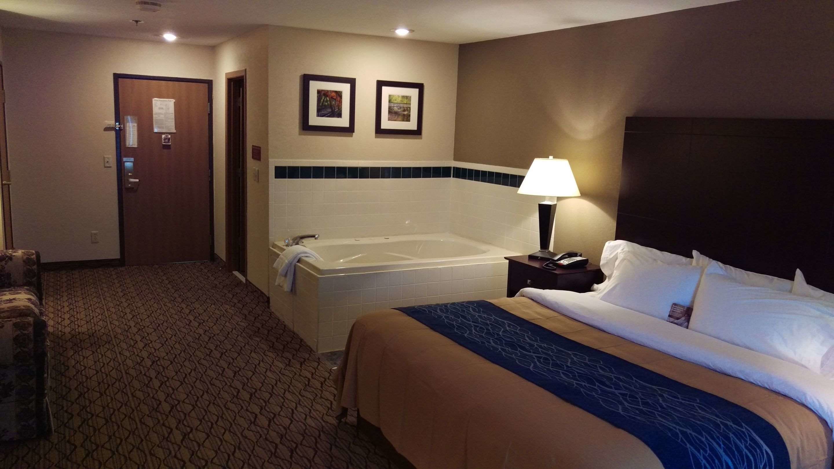 A hotel room with a bed, a bathroom with a bathtub, a nightstand with a lamp, and a door.