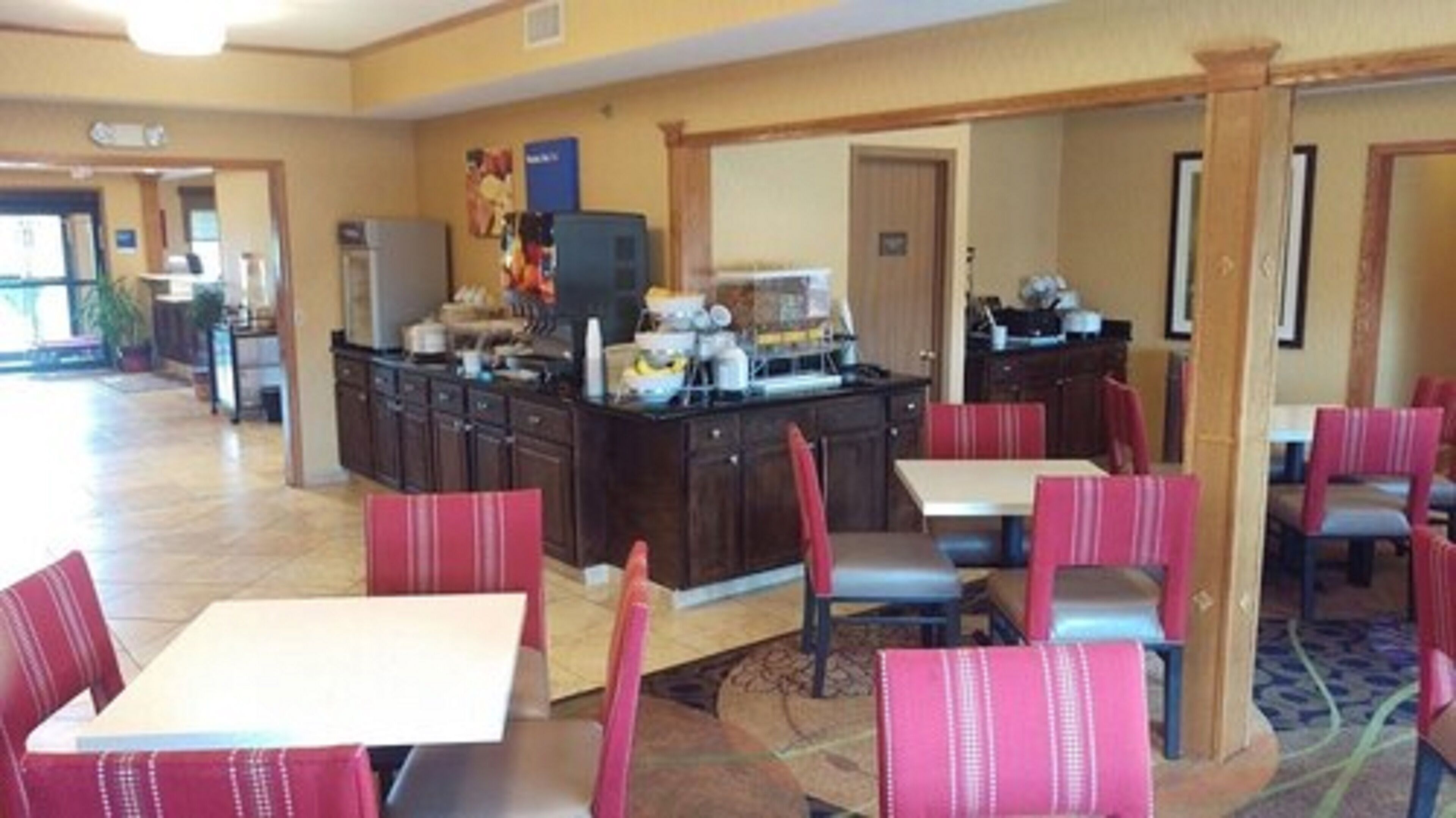 A hotel lobby with a central kitchen area, dining tables with chairs, and a carpeted floor.