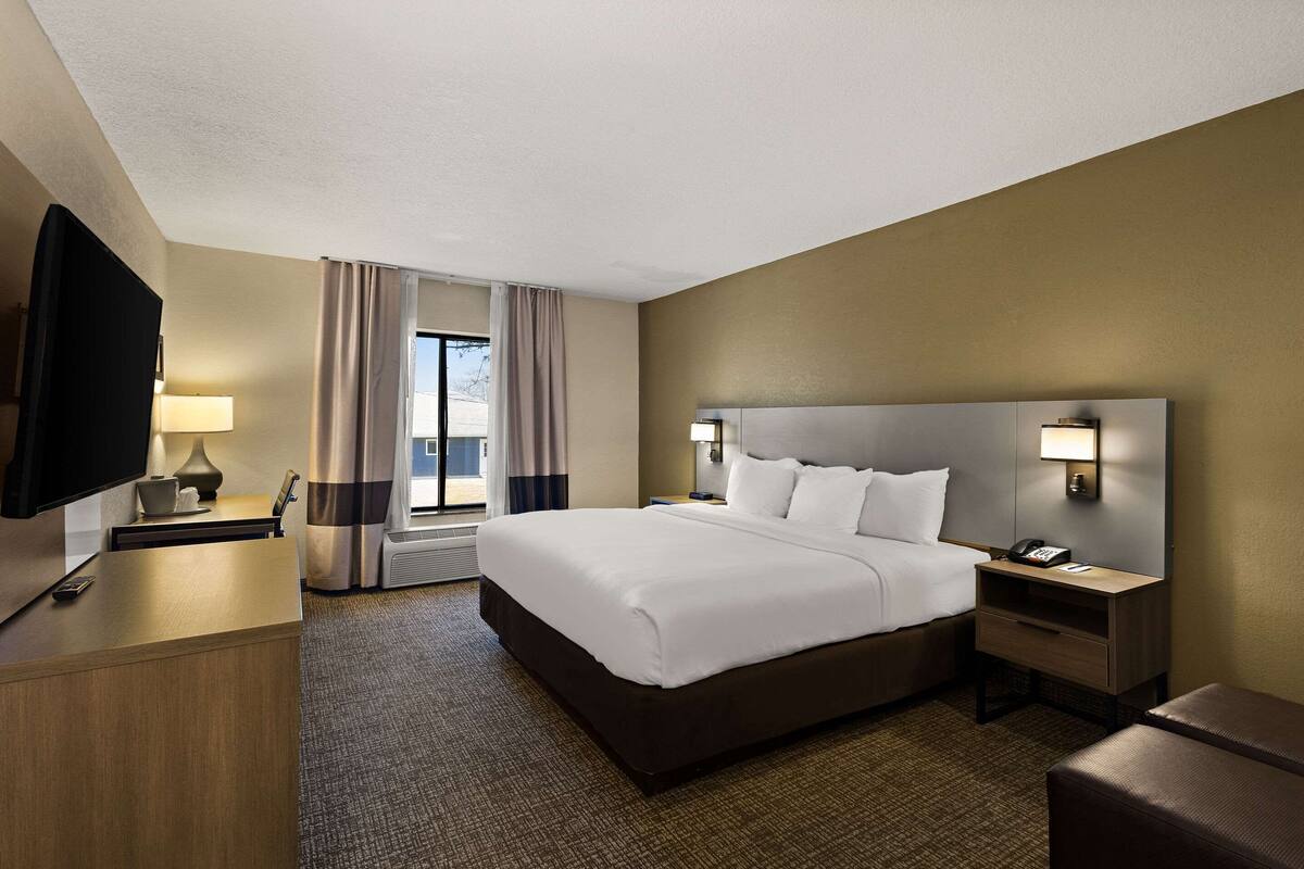 A hotel room with a large bed, a desk with a lamp, a TV, and a nightstand with a phone.
