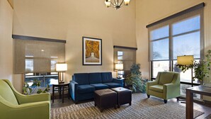 A spacious living room with a blue sofa, green armchair, and a wooden coffee table. There are two lamps, a large window with a view, and a framed picture on the wall. - Comfort Inn Huntington Near University (Huntington)