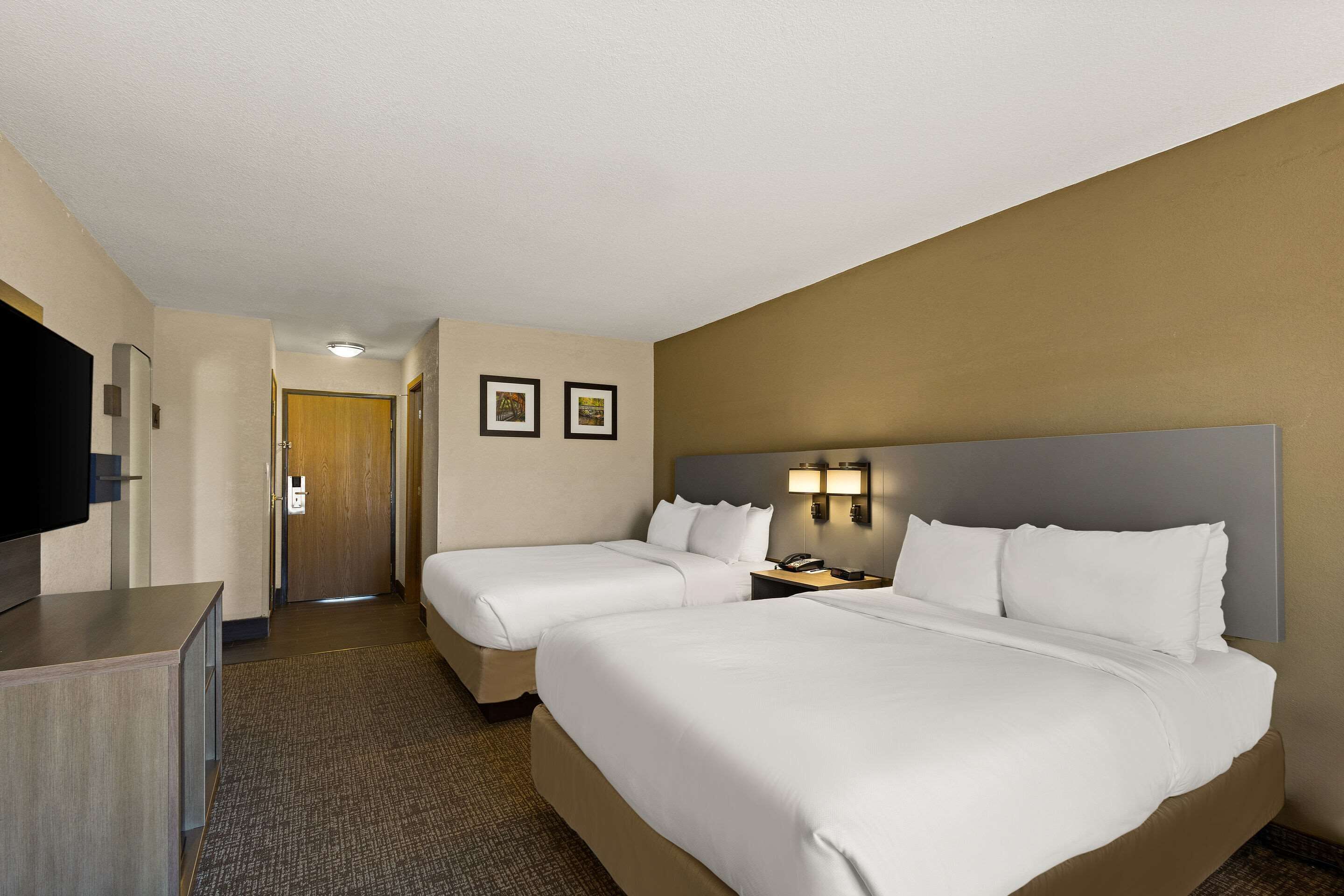A hotel room with two beds, a desk, a TV, and a door leading to a hallway.