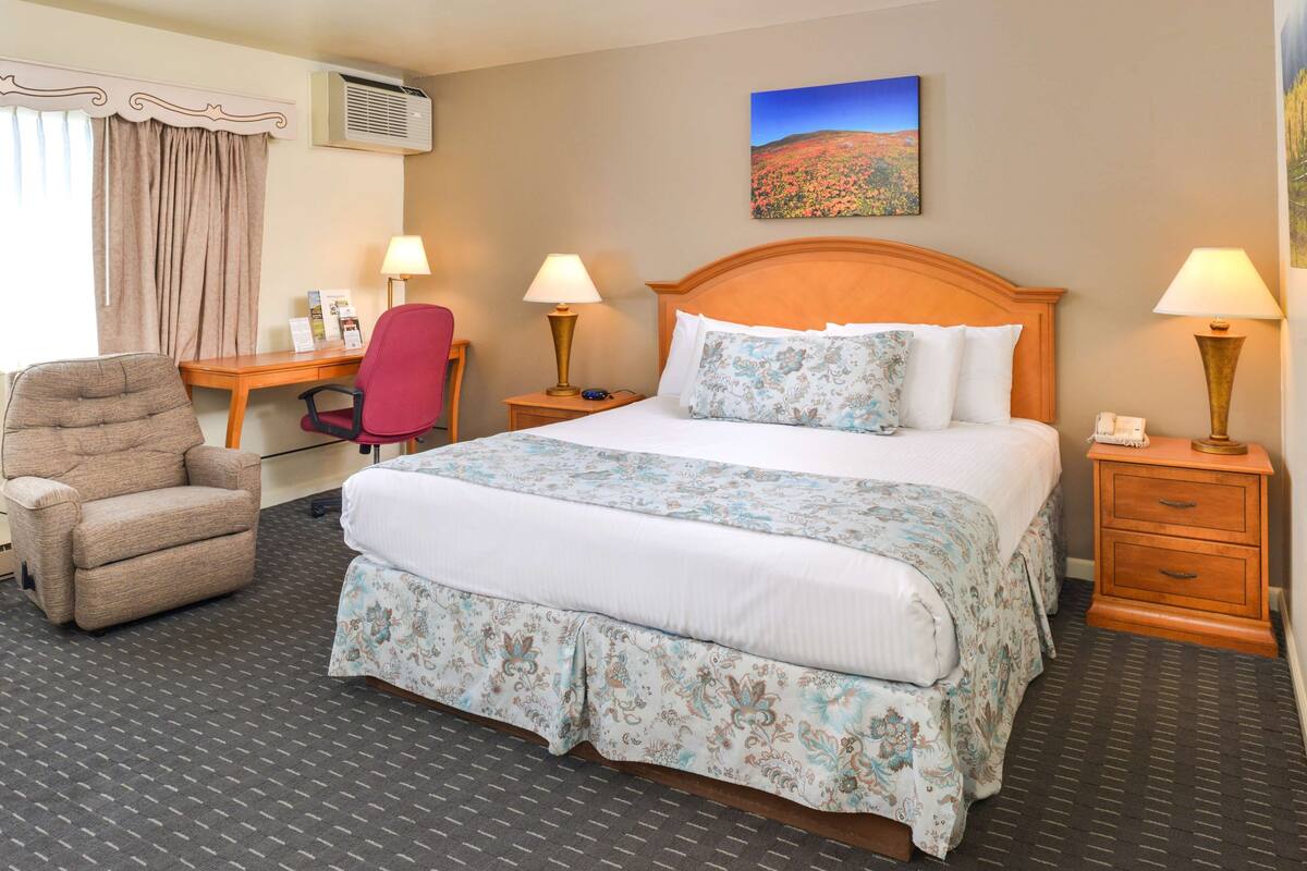 A hotel room with a large bed, two bedside tables with lamps, a desk with a chair, and a window with curtains.