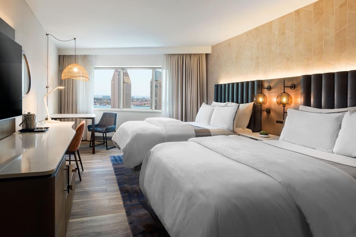 Ein Hotelzimmer mit zwei Betten, einem Schreibtisch, einem Stuhl, einem Fernseher, einem Fenster mit Blick auf die Stadt und einer Wandlampe.