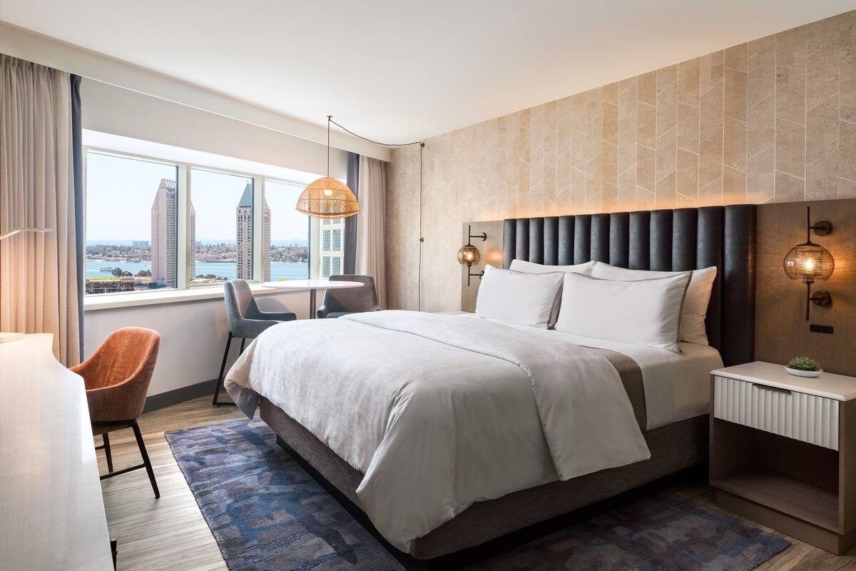 Ein modernes Hotelzimmer mit einem großen Bett, einem Schreibtisch und einem Stuhl. Durch das Fenster ist ein Blick auf Gebäude zu sehen.