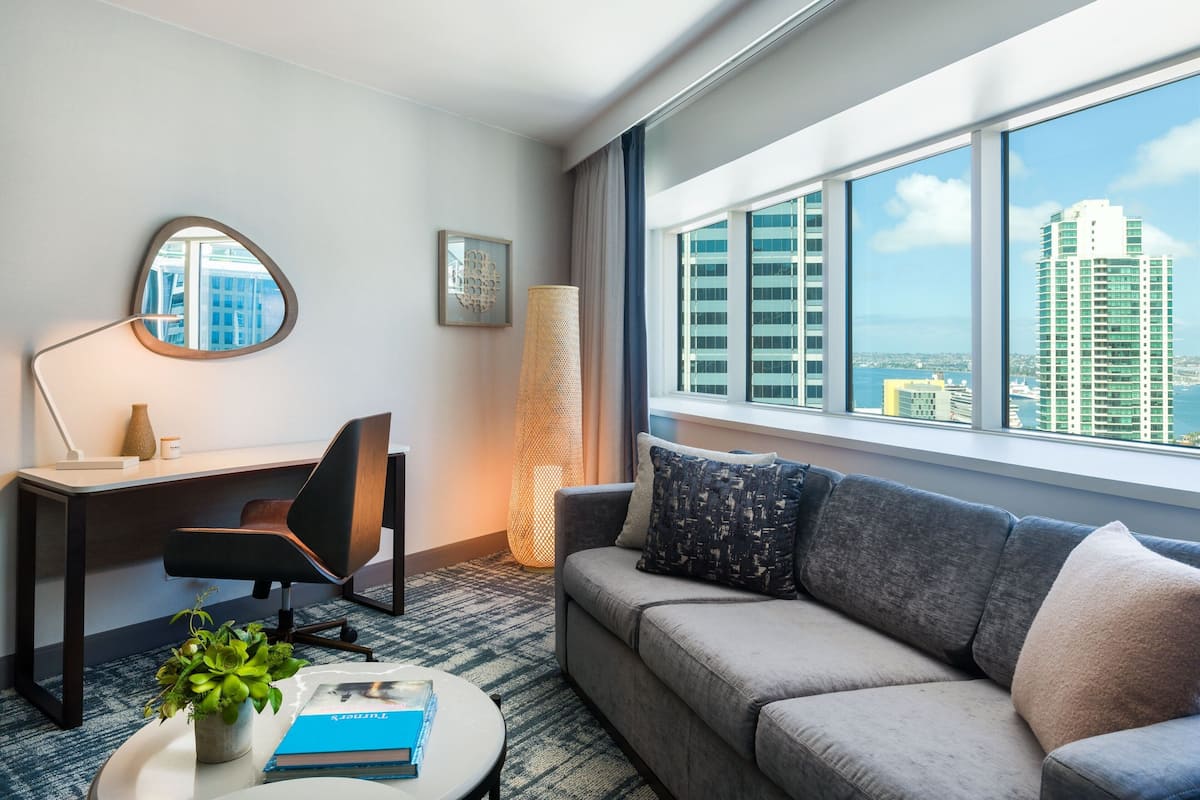 Ein modernes Hotelzimmer mit großem Fenster, einer Couch, einem Schreibtisch mit Stuhl, einer Lampe und einer kleinen Pflanze.