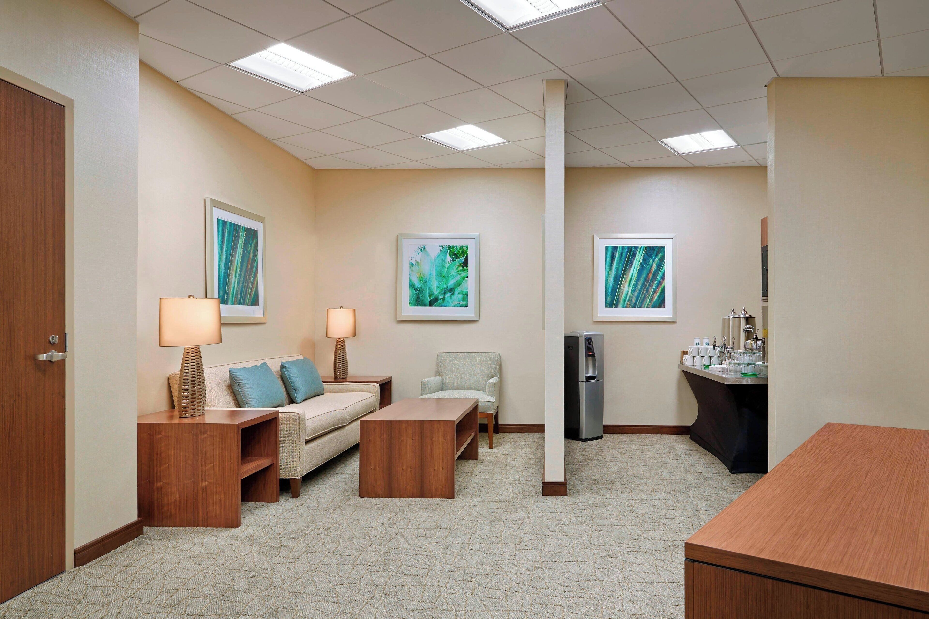A hotel room with a sofa, two armchairs, a wooden coffee table, a desk, a refrigerator, and a small kitchenette.
