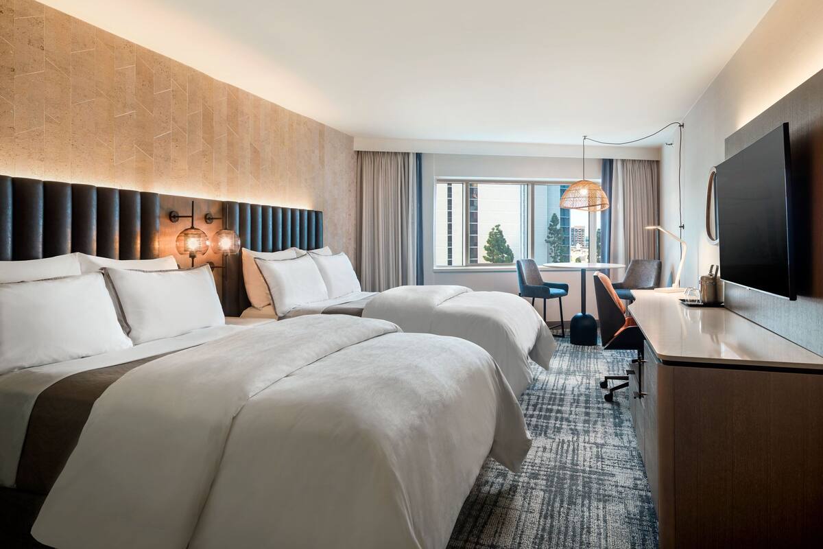 Ein Hotelzimmer mit zwei Betten, einem Schreibtisch, einem Fernseher und einem großen Fenster mit Blick auf ein Gebäude und Bäume.