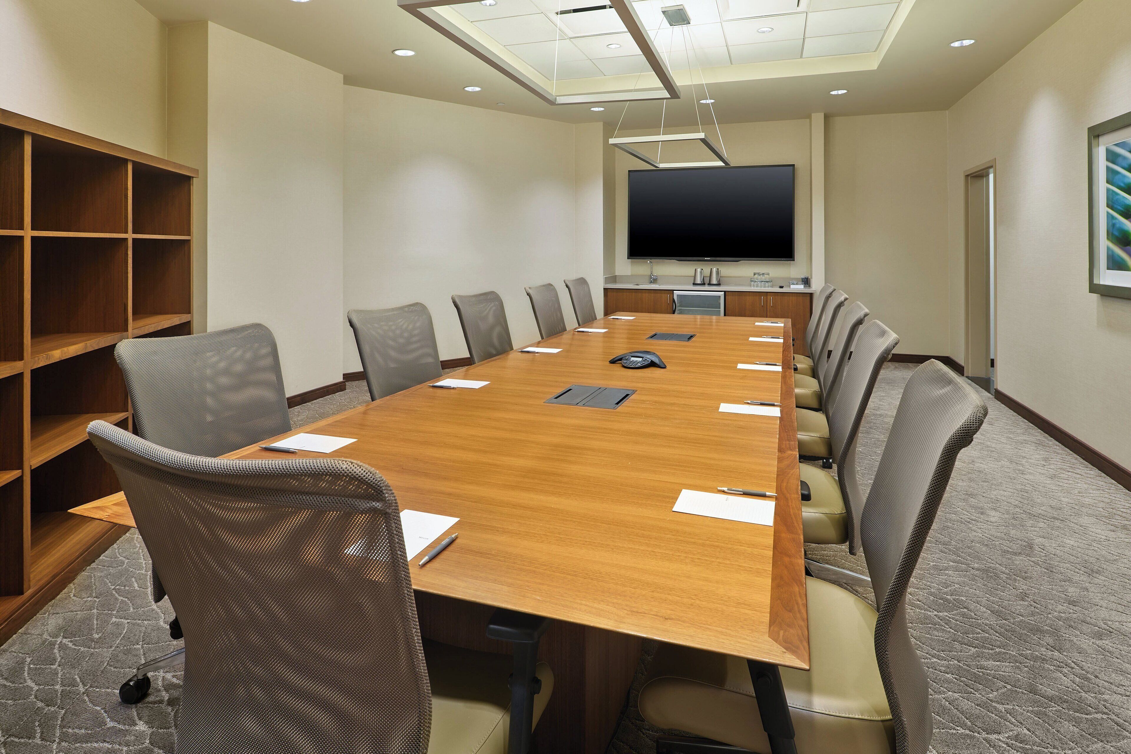 A conference room with a long wooden table, grey mesh chairs, a large flat-screen TV, and a bookshelf.