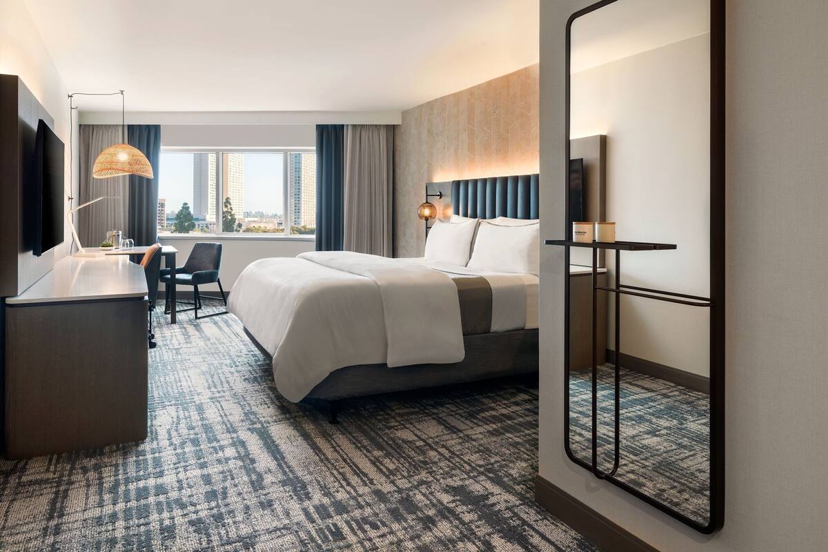 Ein Hotelzimmer mit einem großen Bett, einem Schreibtisch, einem Sessel und einem großen Spiegel, mit Blick auf die Stadt durch das Fenster.