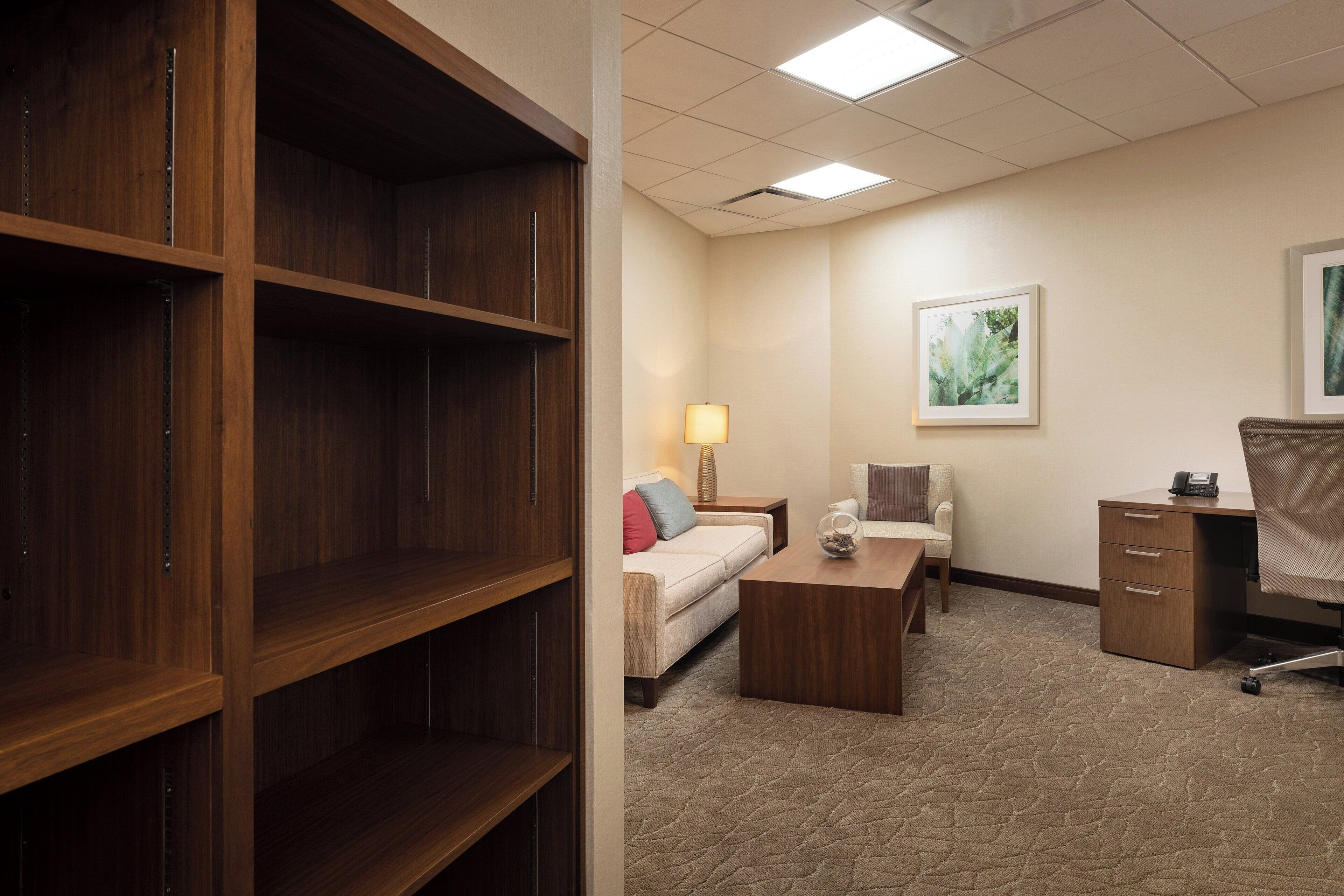 Een hotelkamer met een houten boekenkast, een bank, een salontafel, een bureau met stoel en een lamp.