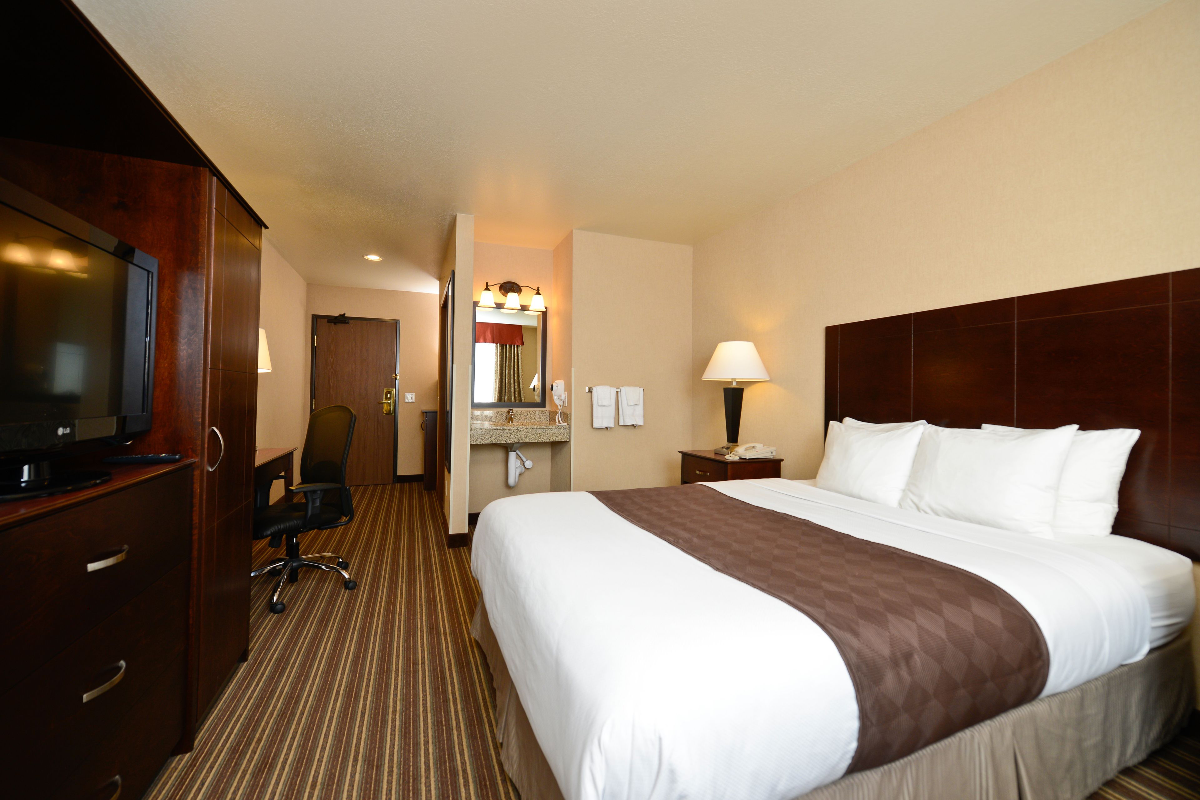 A hotel room with a large bed, a desk with a television, a chair, a bathroom with a mirror and lights, and a nightstand with a lamp.