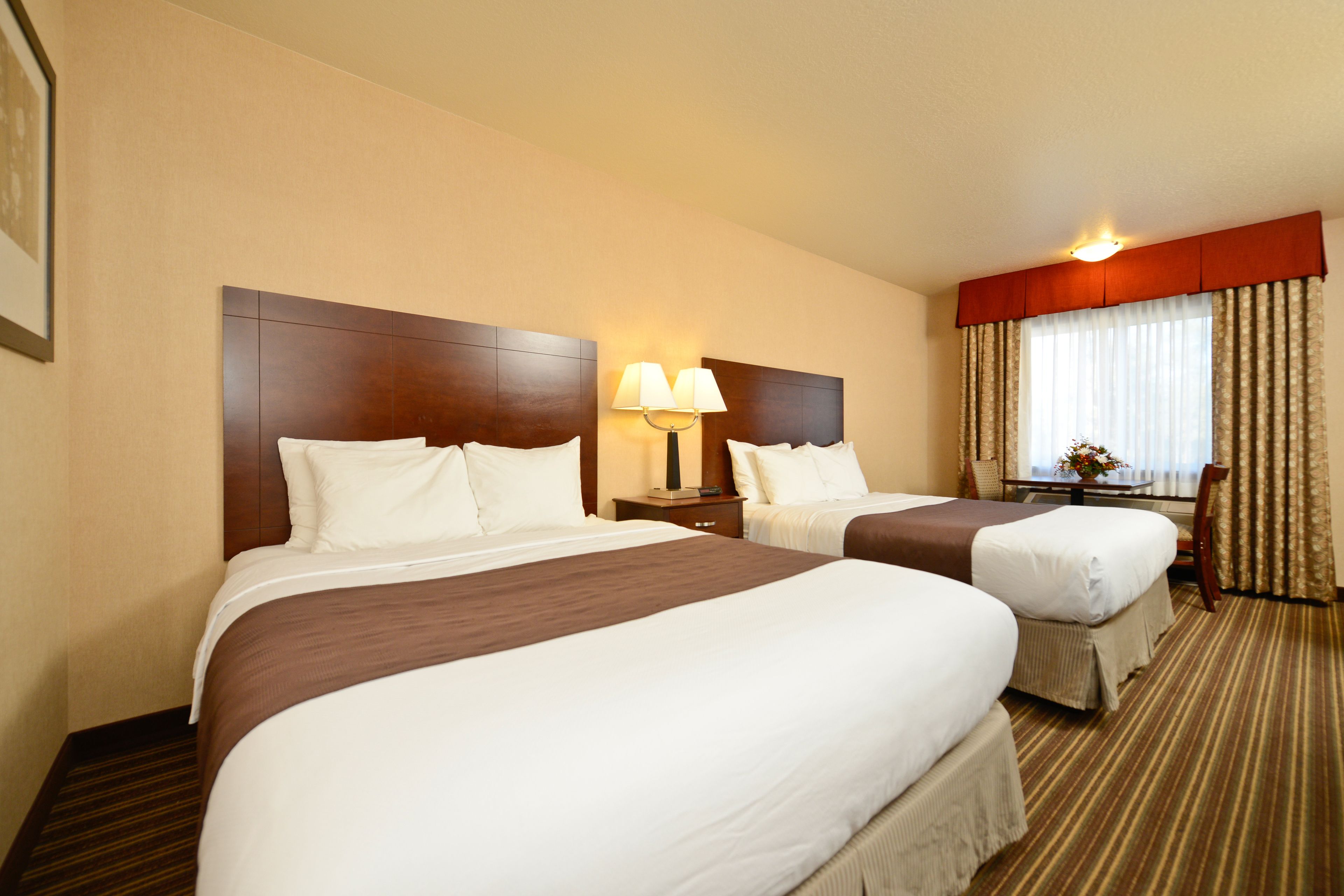 A hotel room with two beds, a nightstand with a lamp, a window with curtains, and a carpeted floor.