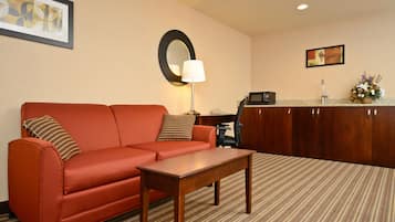 Habitación de hotel con un sofá rojo, una mesa de centro de madera, un escritorio con una computadora, un espejo y un cuadro en la pared.