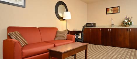 A hotel room with a red sofa, a wooden coffee table, a desk with a computer, a mirror, and a framed picture on the wall.