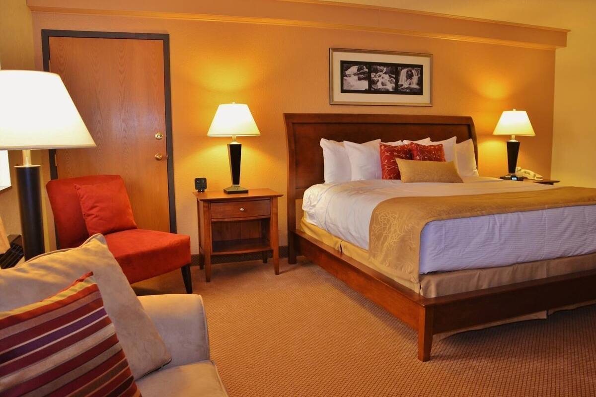 A hotel room with a bed, two bedside tables with lamps, a chair, a desk, and a framed picture on the wall.