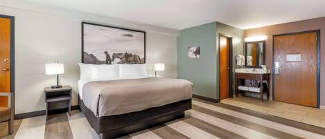 A hotel room with a large bed, bedside tables with lamps, a bathroom with a mirror, and a wooden floor.