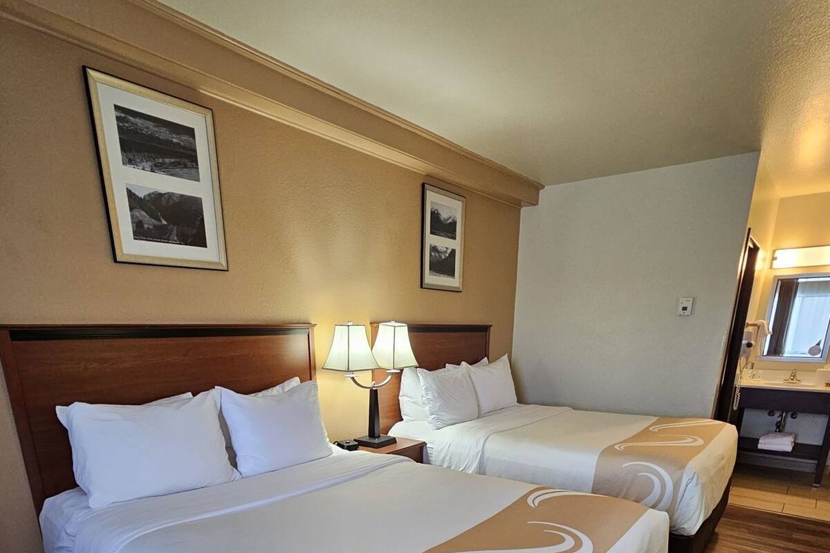 A hotel room with two beds, wooden flooring, a nightstand with lamps, and framed pictures on the wall.