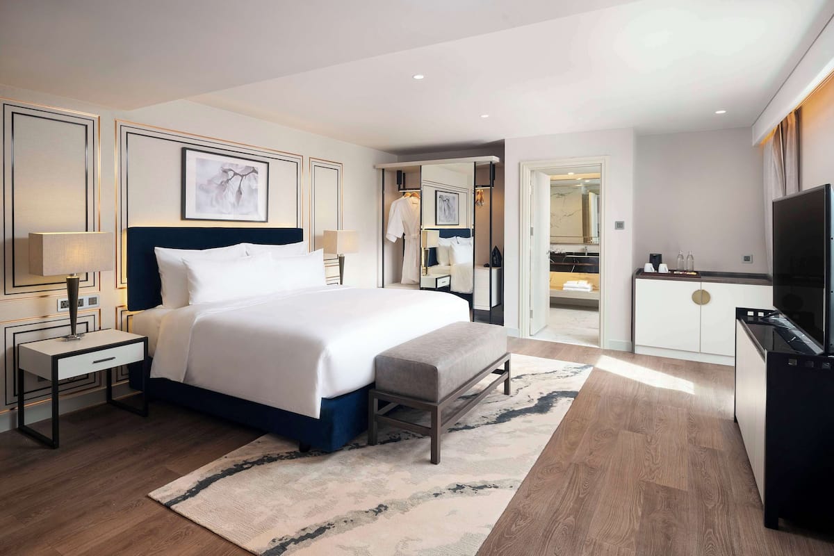 Una habitación de hotel moderna con una cama grande, mesitas de noche, un banco, un televisor y un baño visible a través de una puerta abierta.