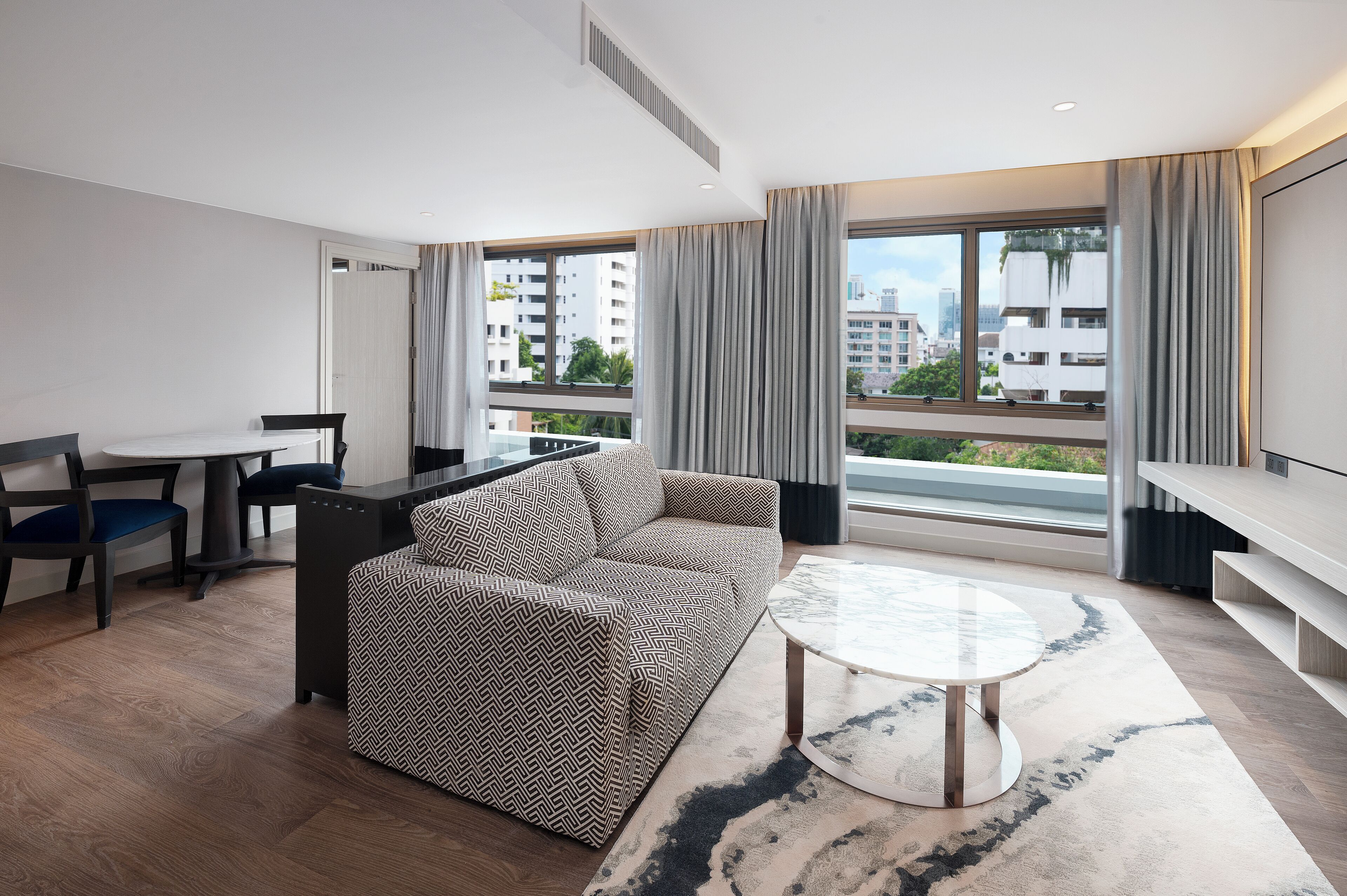 A modern hotel room with a sofa, dining table, and a large window offering a city view.