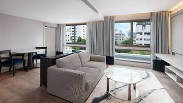 A modern hotel room with a sofa, dining table, and a large window offering a city view.
