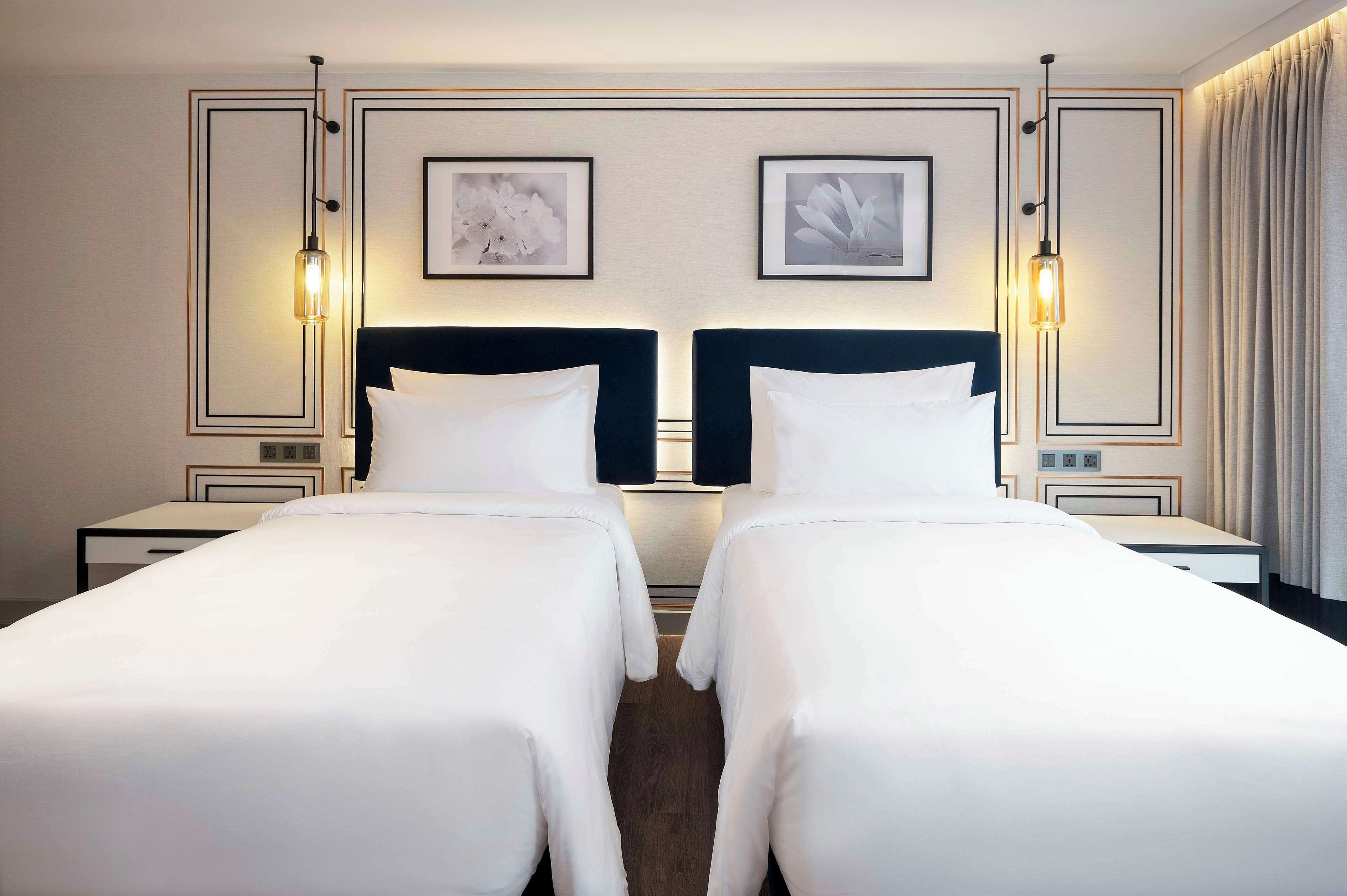 Quarto de hotel com duas camas, cabeceira escura, roupa de cama branca, obras de arte emolduradas e luminária.