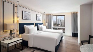 Quarto de hotel com duas camas, uma escrivaninha, uma cadeira, uma janela grande com vista para a cidade e um tapete estampado.