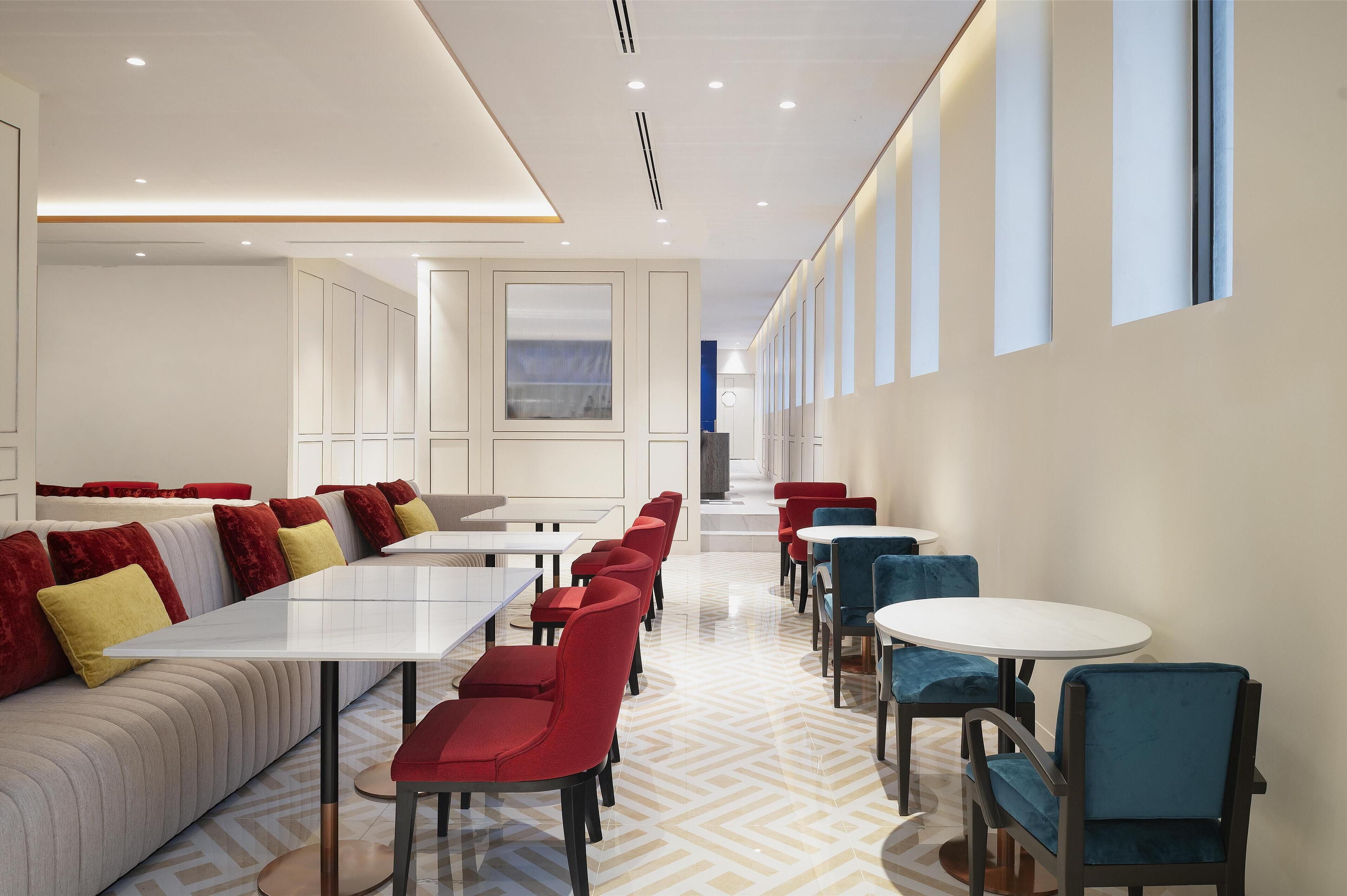 A modern hotel lobby with a long reception desk, seating area with red and blue chairs, and a patterned floor.