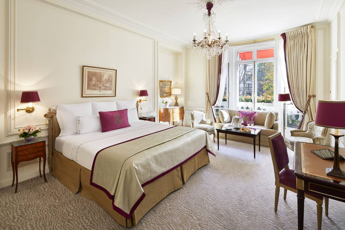 Hotelkamer met een groot bed, zithoek met een tafel en stoelen, een kroonluchter en uitzicht op het groen.