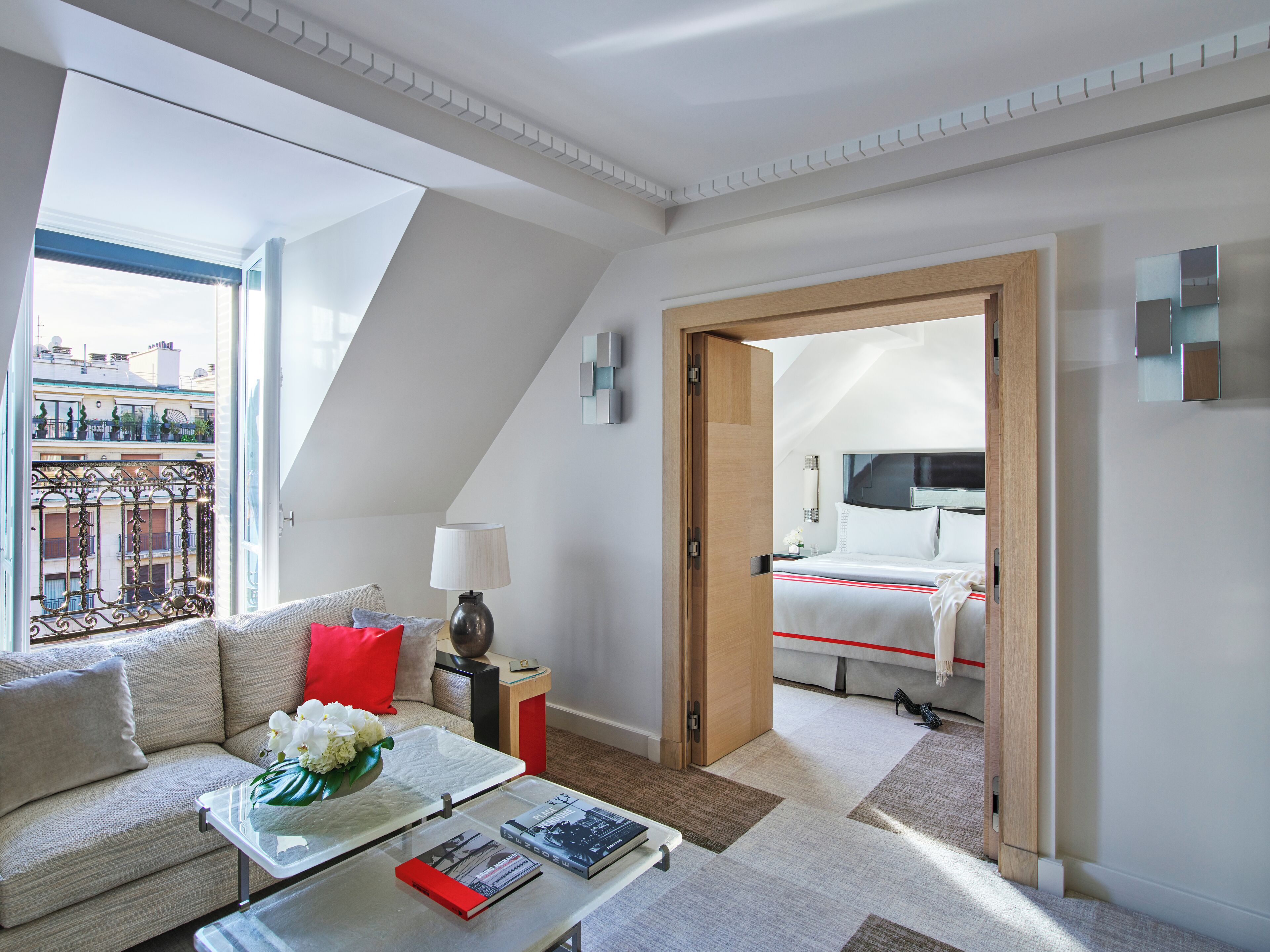 A modern hotel room with a balcony, a sofa, a coffee table with books, and a bed with a red-striped bedspread.