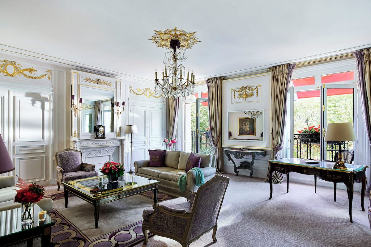 Een klassieke, elegant ingerichte woonkamer met een kroonluchter, comfortabele zithoeken en uitzicht door grote ramen.