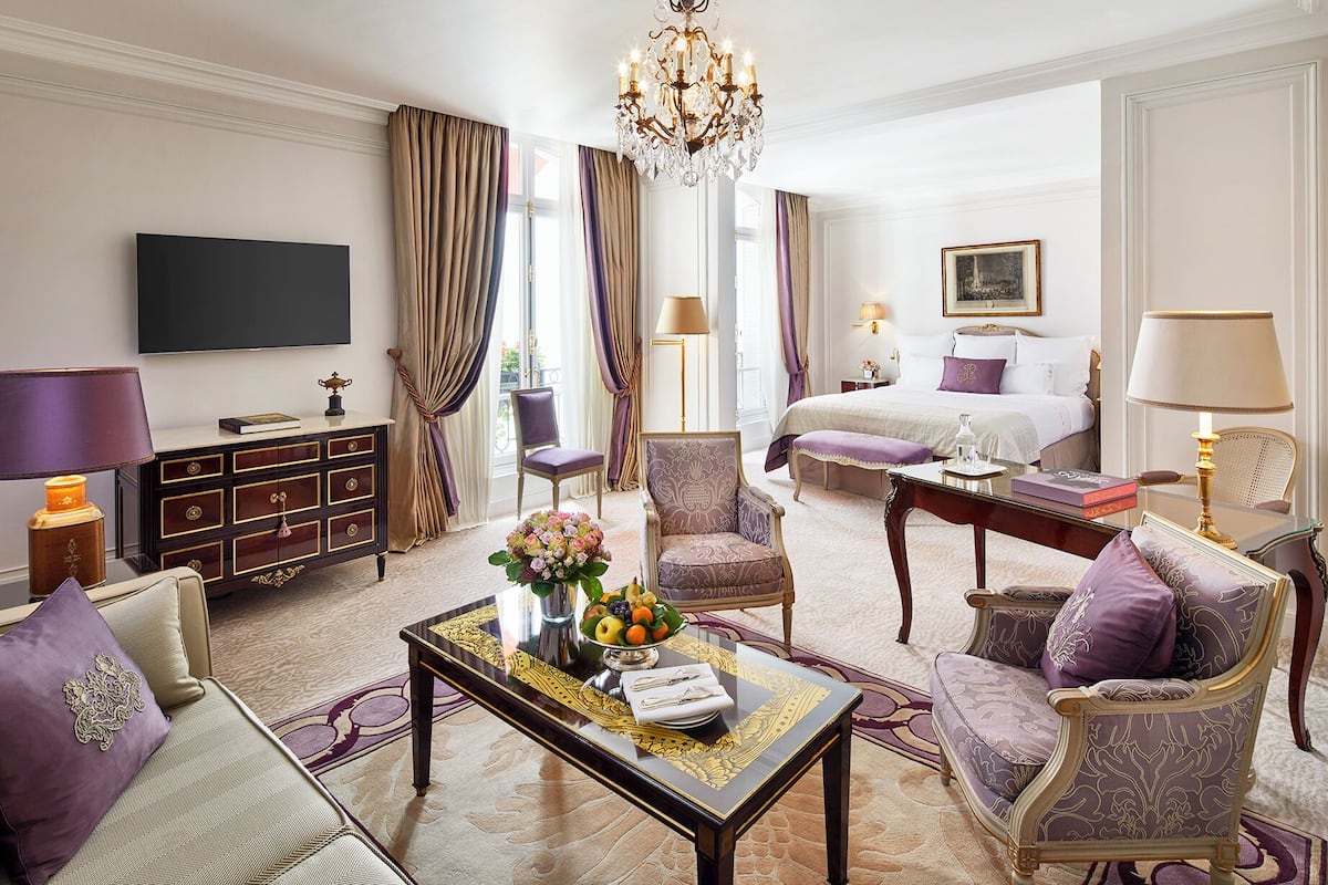 A hotel room with a bed, a TV, a dining table with chairs, a sofa, and a chandelier.