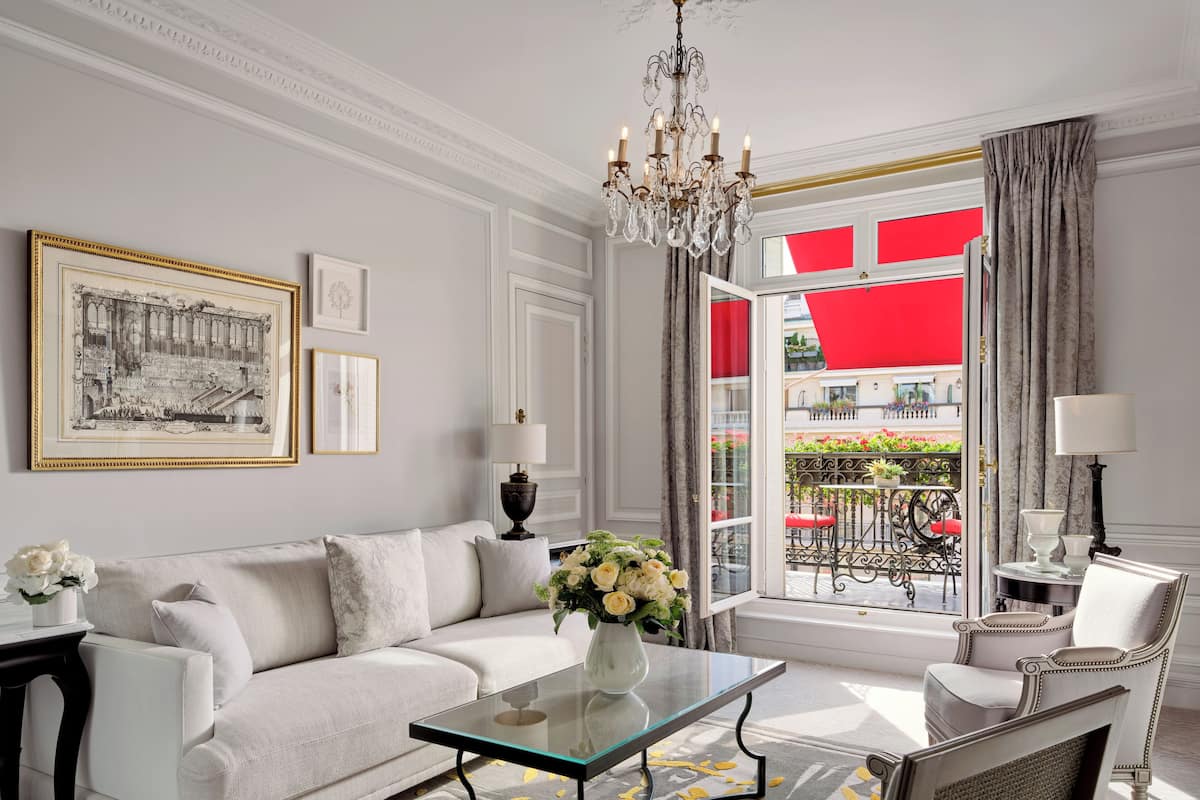 Een moderne woonkamer met een witte bank, een glazen salontafel en een kroonluchter. Er is uitzicht op een rode luifel en een balkon met tuinmeubilair.