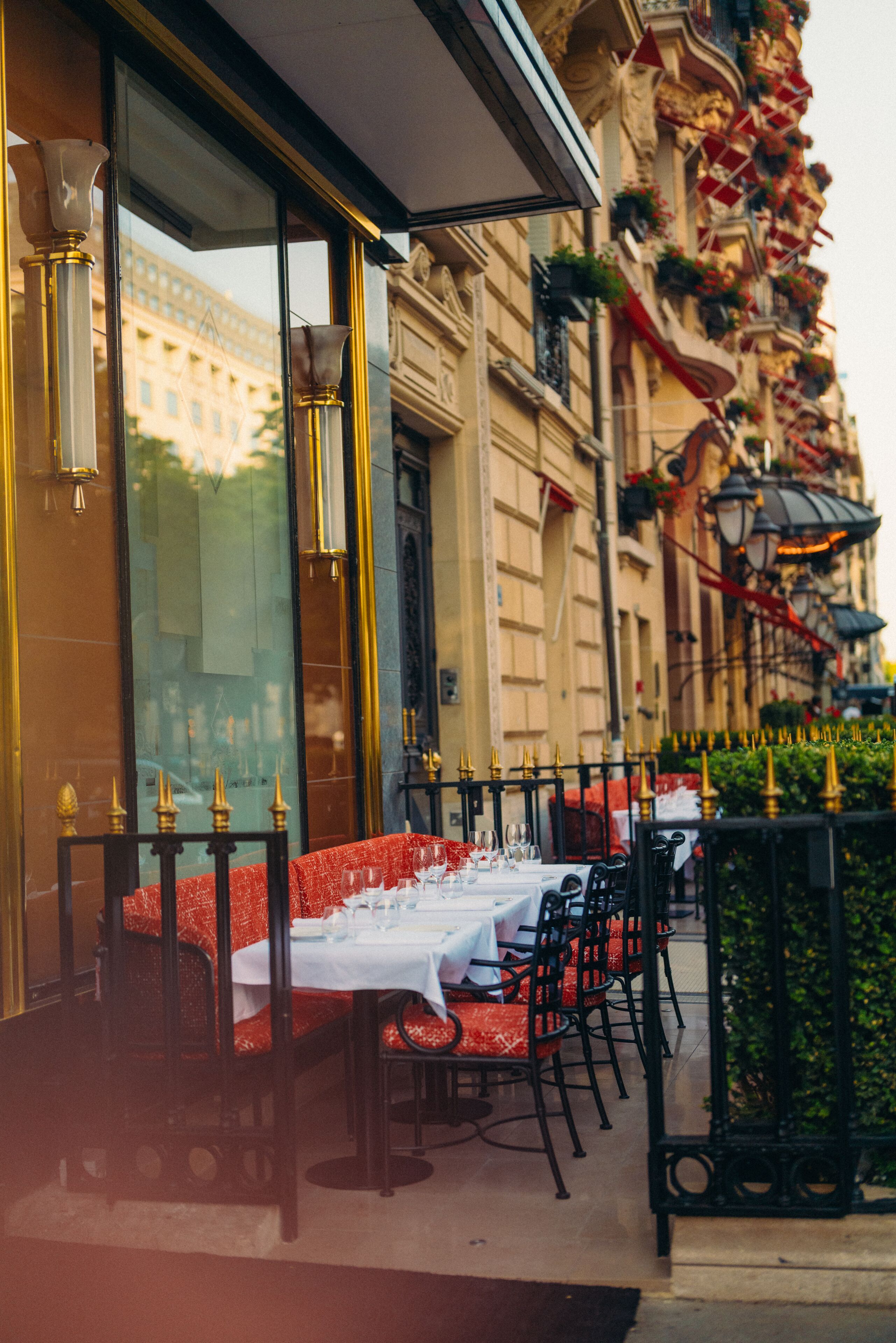 An outdoor dining area with tables set for two, featuring red and white tablecloths and black chairs.