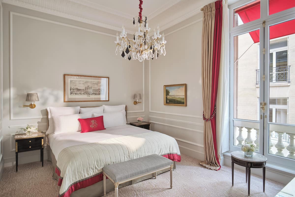 Een hotelkamer met een groot bed, een bankje, een kroonluchter, een raam met rode gordijnen en twee schilderijen aan de muur.