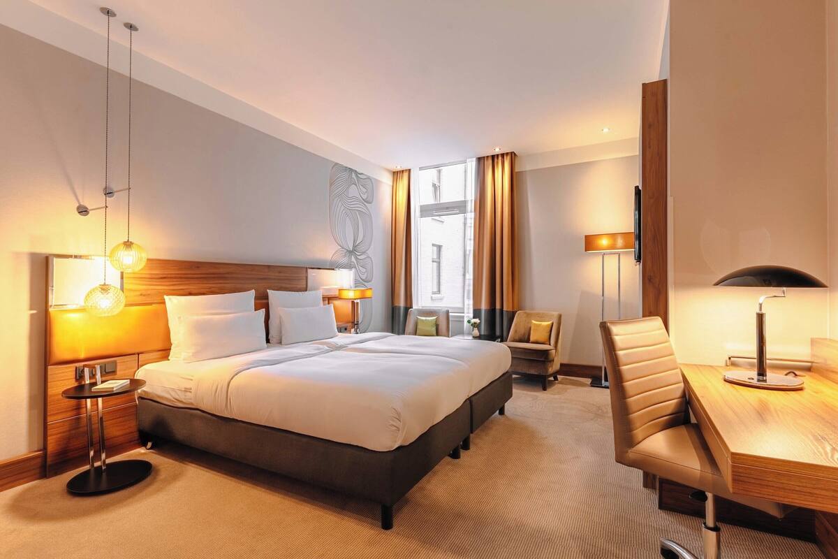 Une chambre d’hôtel avec un grand lit, des tables de chevet avec des lampes, un bureau avec une chaise et une grande fenêtre avec des rideaux.