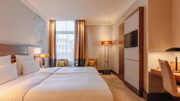 Ein Hotelzimmer mit einem großen Bett, einem Schreibtisch, einem Sessel, einem Fernseher und einem Fenster mit Vorhängen.