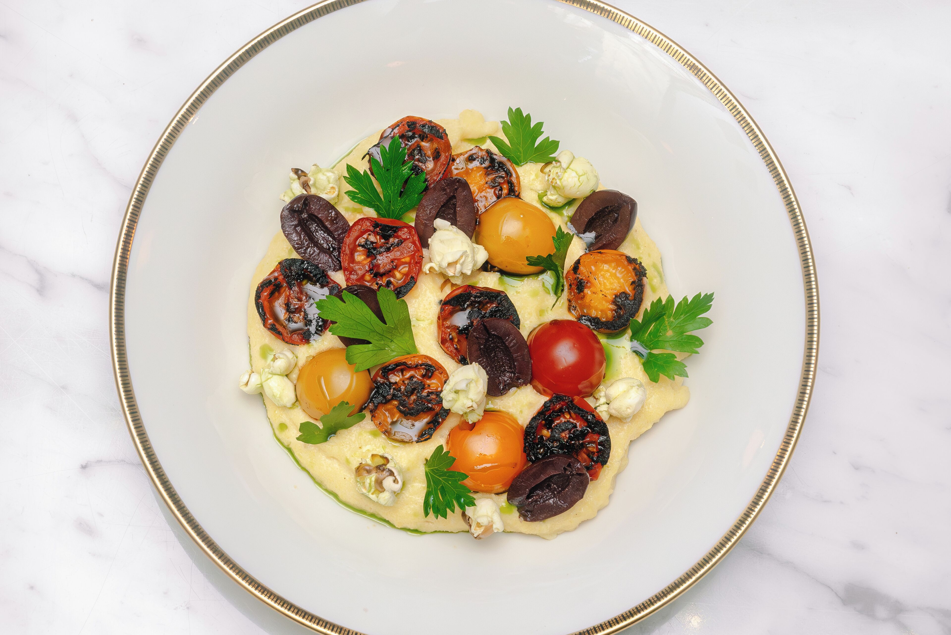 Ein weißer Teller mit goldener Kante, gefüllt mit einem Salat aus Cocktailtomaten, Oliven und Blattsalaten.