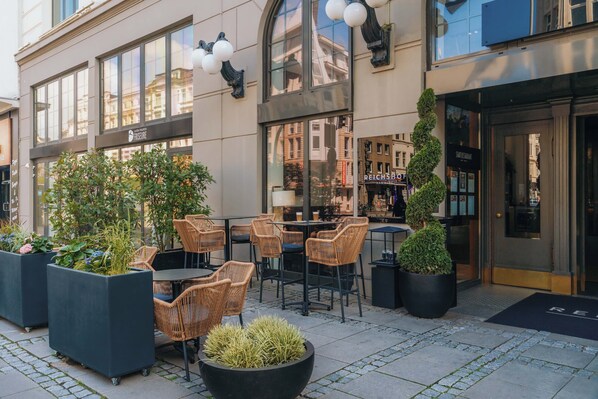 Un espace extérieur aménagé avec des chaises et des tables en osier, des plantes en pot et une façade de bâtiment moderne dotée de grandes fenêtres et d’une enseigne de restaurant.