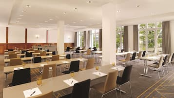 Un salón de conferencias con varias mesas y sillas, ventanas amplias y un techo alto con iluminación empotrada.