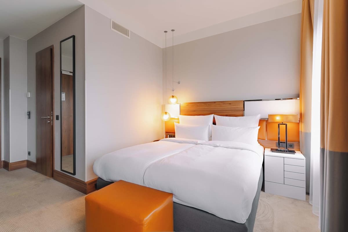 Une chambre d’hôtel avec un grand lit, une table de chevet avec une lampe, une tête de lit en bois et un petit banc orange.