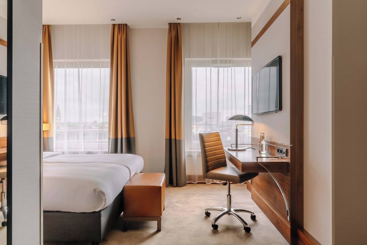 Une chambre d’hôtel équipée d’un lit, d’un bureau, d’une chaise, d’une télévision et de grandes fenêtres avec des rideaux.