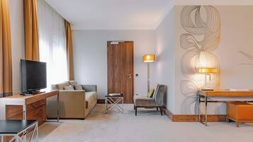 A modern hotel room with a wooden desk, a television, a sofa, a chair, and a lamp.