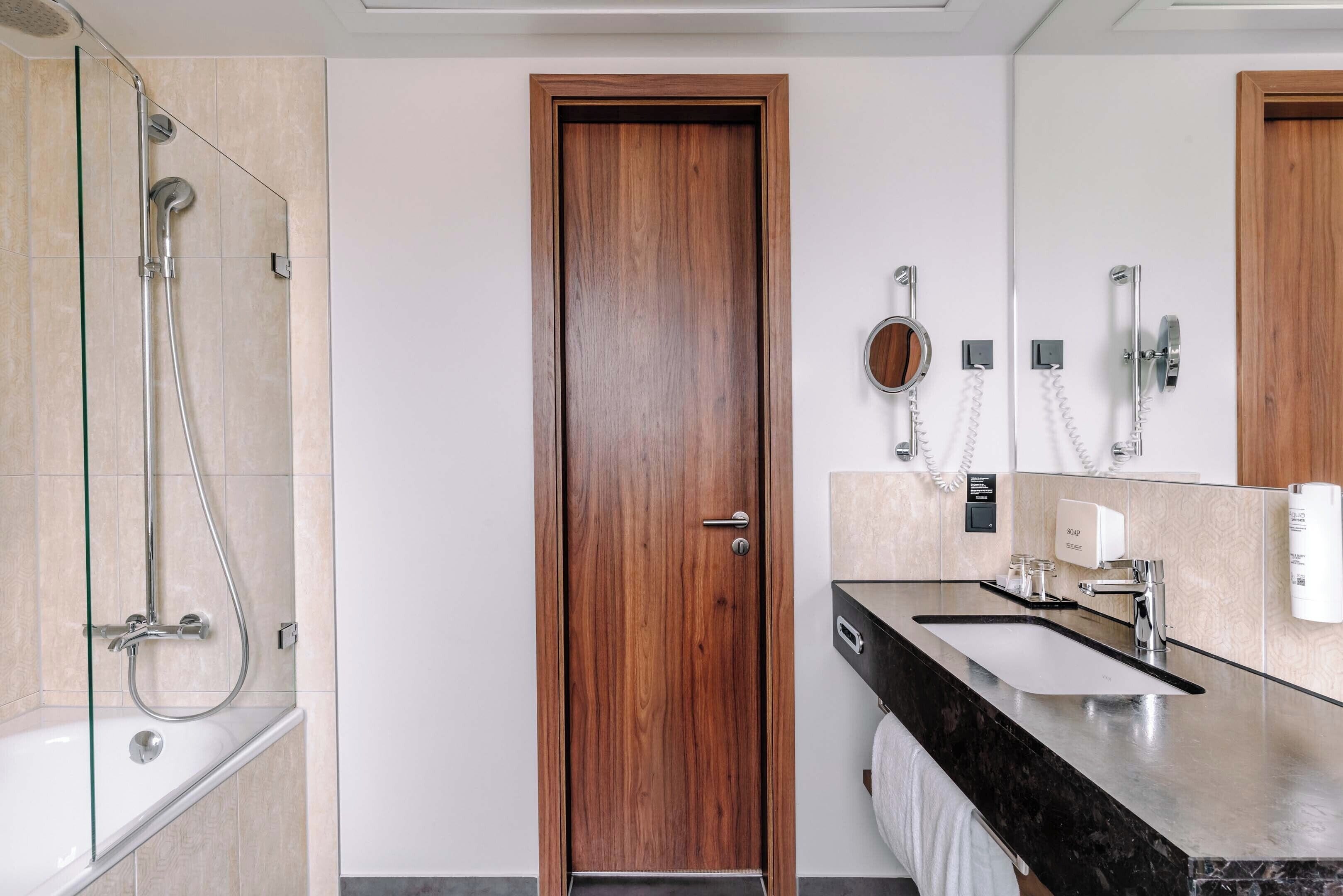 Ein modernes Badezimmer mit Holztür, Duschkabine aus Glas, Waschbecken mit schwarzer Arbeitsplatte und Spiegel.