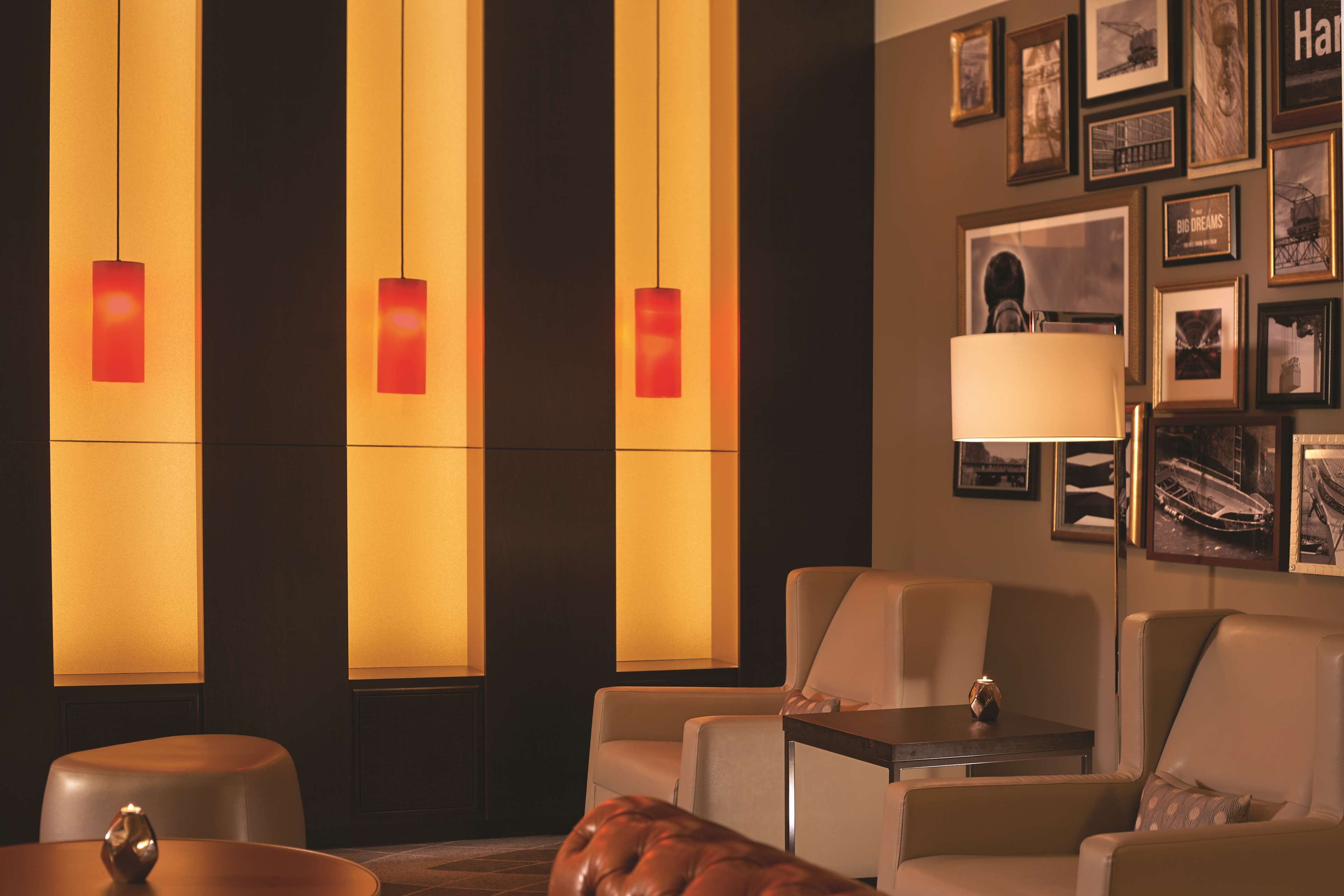 A hotel room with a seating area featuring a sofa, a chair, and a small table. There are wall-mounted lamps with red shades and framed pictures on the wall.