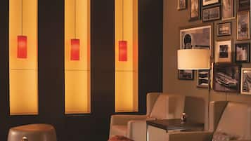 Quarto de hotel com área de estar, incluindo um sofá, uma poltrona e uma mesinha. Há luminárias fixadas na parede com abajures vermelhos e quadros enquadrados.
