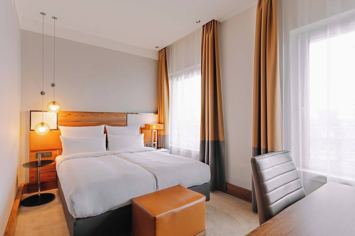 Une chambre d’hôtel avec un grand lit, deux tables de chevet avec des lampes, une tête de lit en bois, un fauteuil en cuir et une grande fenêtre avec des rideaux.