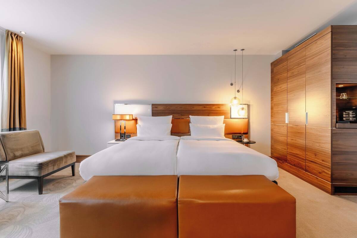 Une chambre d’hôtel avec un grand lit, deux tables de chevet avec des lampes, une armoire en bois, un fauteuil en cuir et une grande fenêtre avec des rideaux.