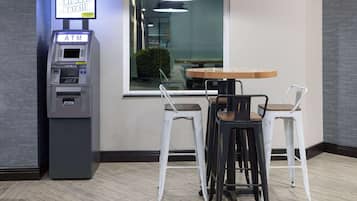 Een pinautomaat, een houten tafel en twee witte barstoelen in een modern interieur.